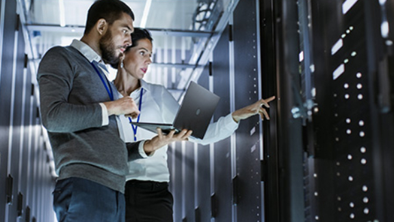 A man holding a laptop standing next to a woman pointing at a control panel