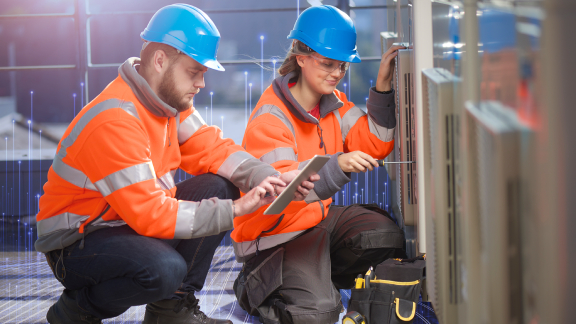 Two maintenance workers kneeling before a device, while one scrolls on a tablet