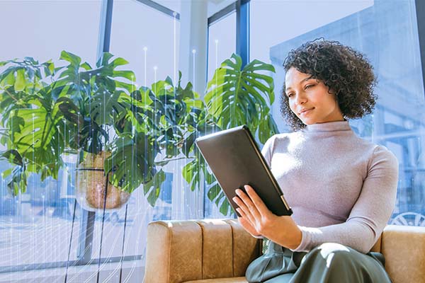 A woman sitting on a couch near plants and looking at a tablet, with OpenBlue graphics in the background